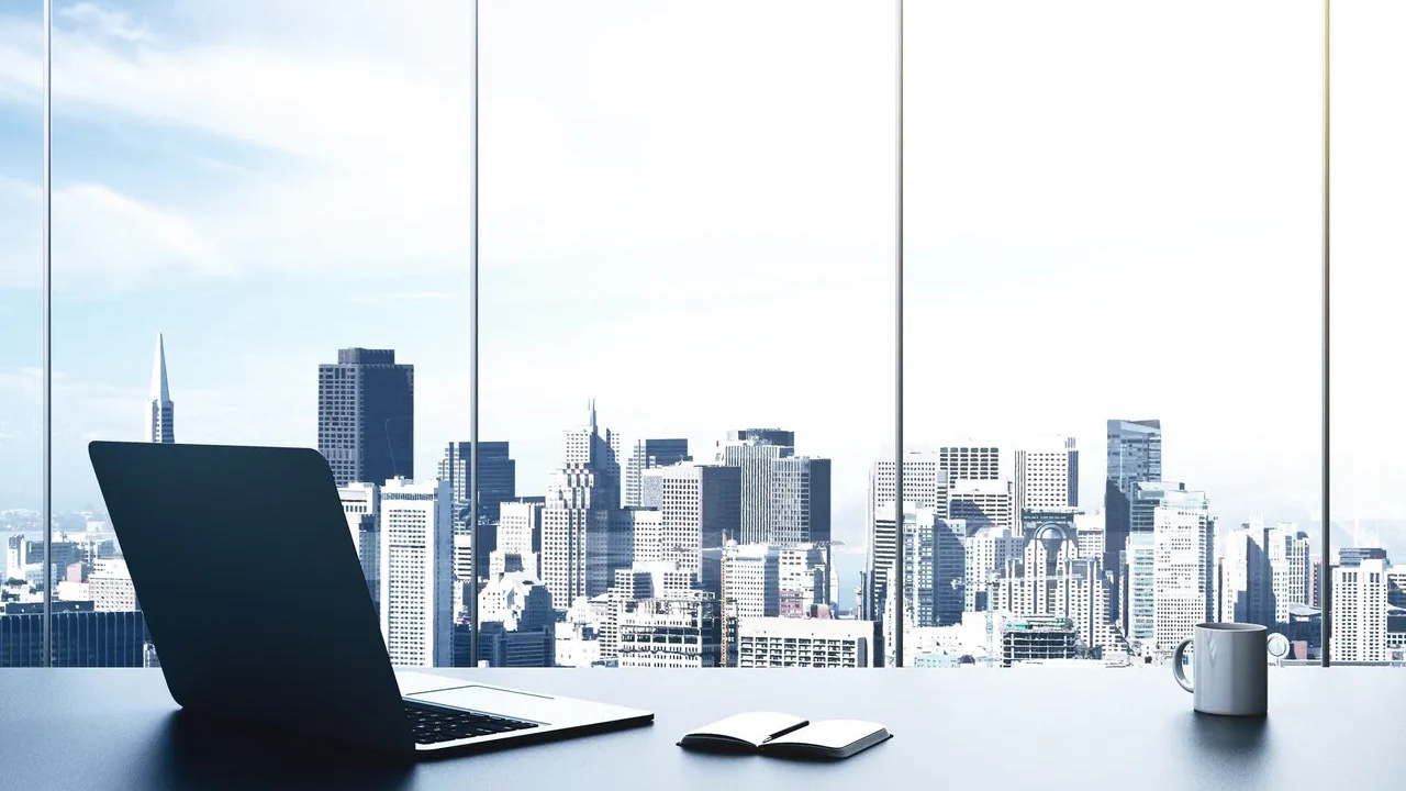 A modern office desk with an open laptop, a notebook, and a coffee mug, set in front of large windows overlooking a city skyline with tall buildings under a bright sky.