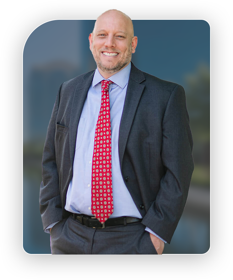A man wearing a dark suit, light blue shirt, and red patterned tie stands smiling with his hands in his pockets in front of a blurred outdoor background.